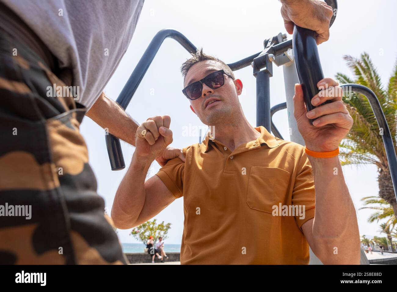 A man assists another in using outdoor exercise equipment on a sunny ...