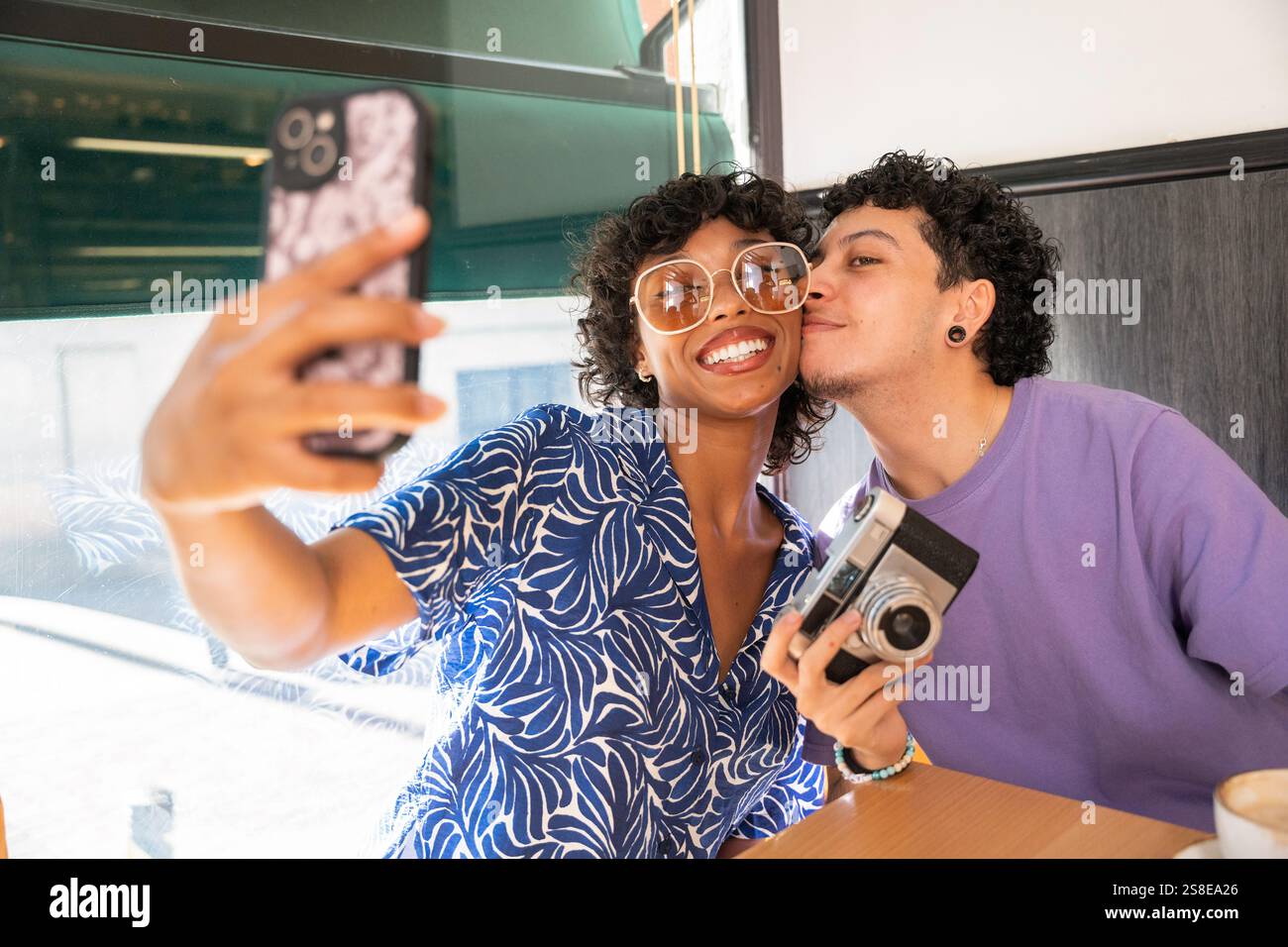 A joyful multiethnic LGBTQ+ couple captures a moment at a cafe. They smile while taking a selfie, showing love and happiness. The atmosphere is warm a Stock Photo
