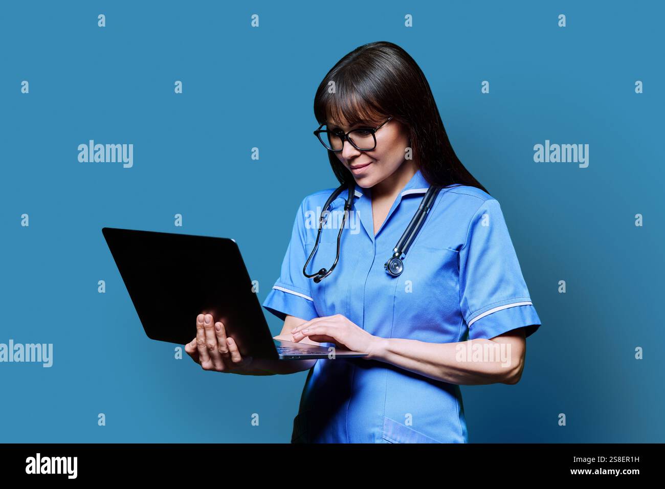 Smiling female nurse using laptop computer, profile view on blue studio ...