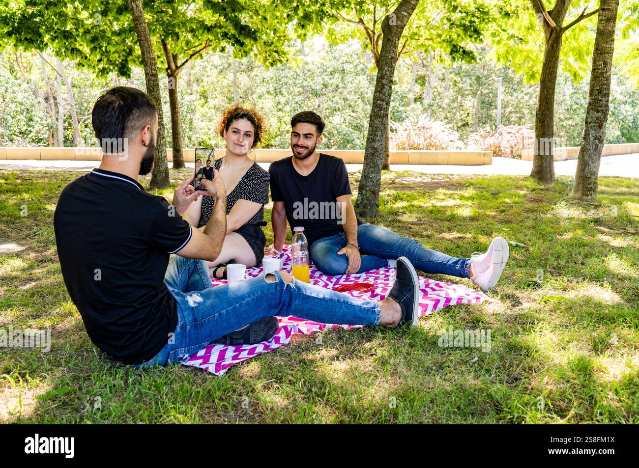 Three young adults enjoy a relaxing picnic on a vibrant blanket in the park, with one man photographing two smiling friends Stock Photo