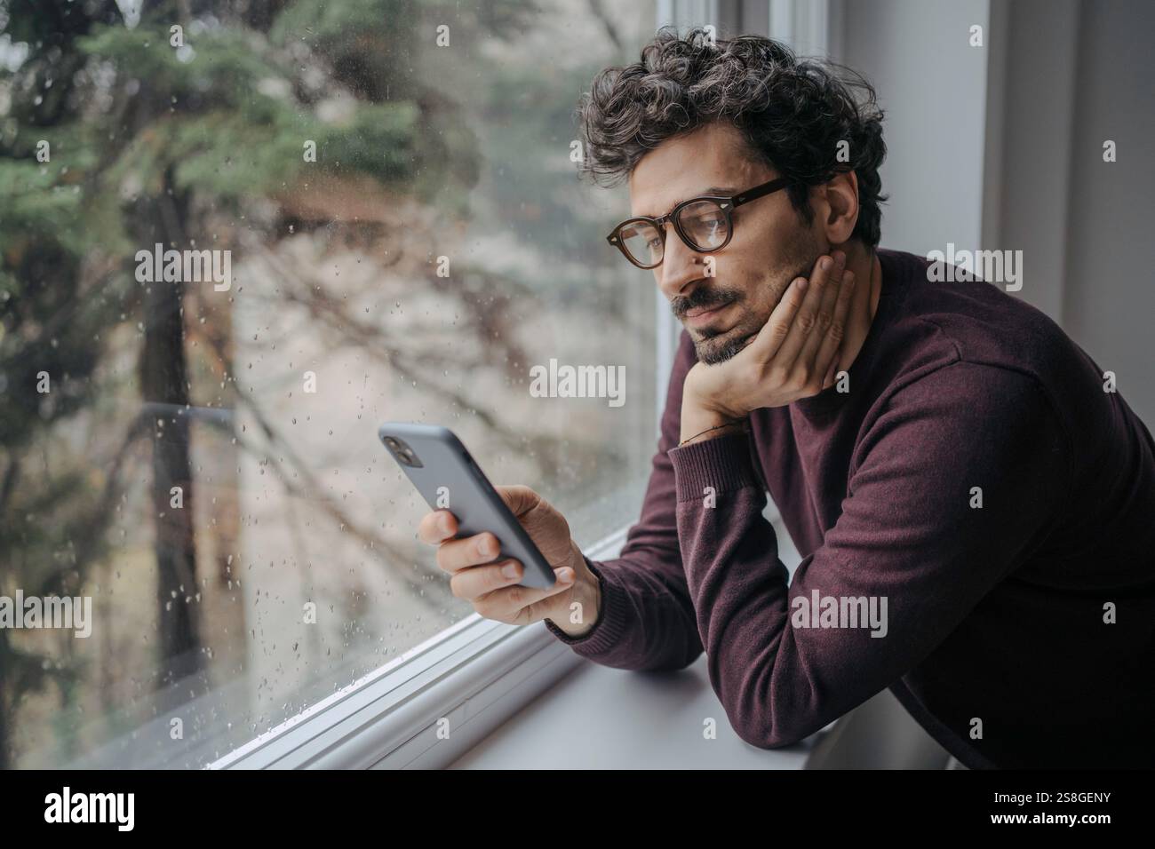 Adult middle eastern good looking man with glasses is scrolling on mobile phone in the morning standing near window. Stock Photo