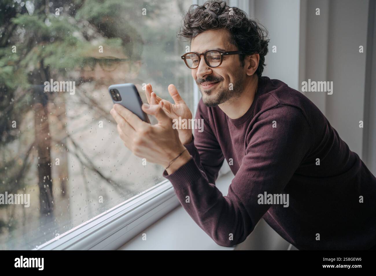 Young handsome middle eastern man with glasses having a video call on mobile phone while standing near window. Stock Photo