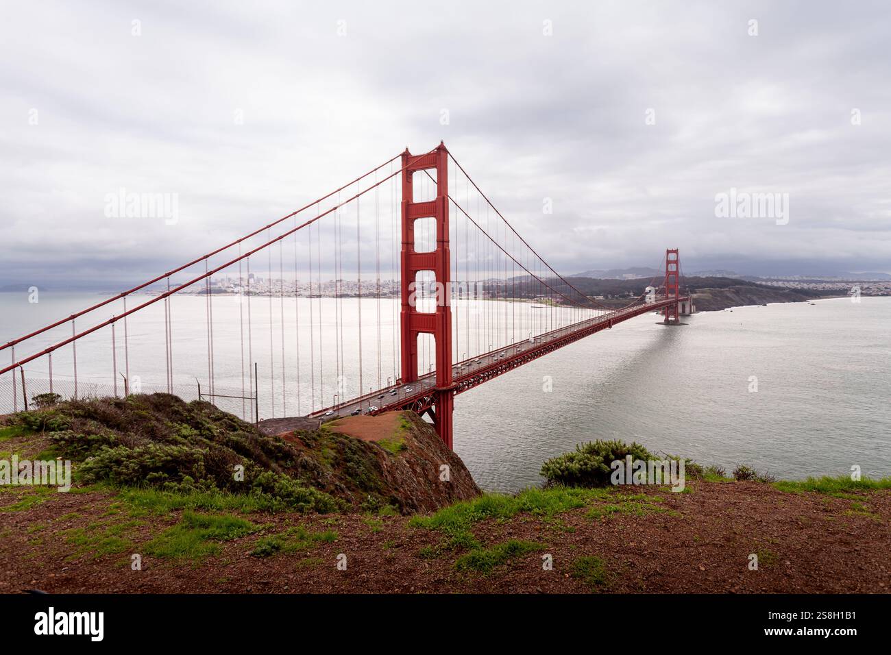 The Golden Gate Bridge stands tall over the bay, surrounded by lush green hills. Soft clouds hang in the sky, reflecting the calm water below. This vi Stock Photo