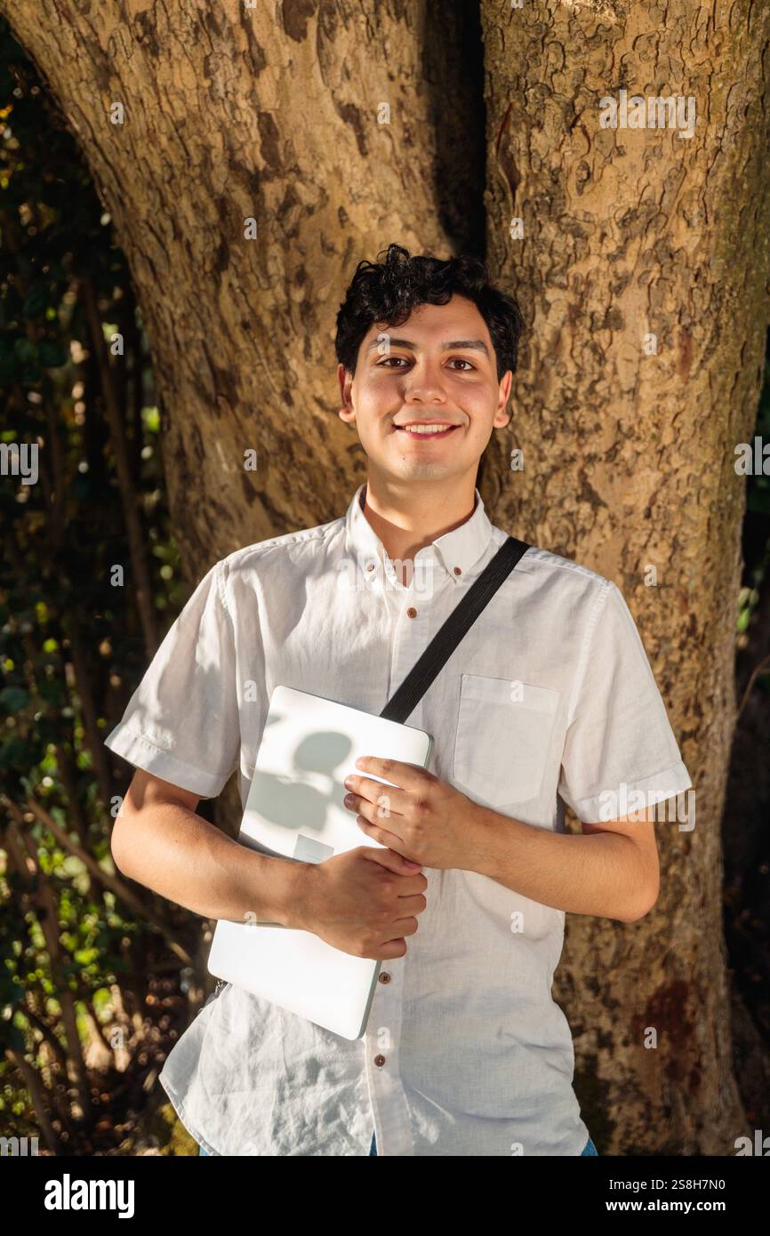 Smiling student holding laptop leaning against tree in park Stock Photo