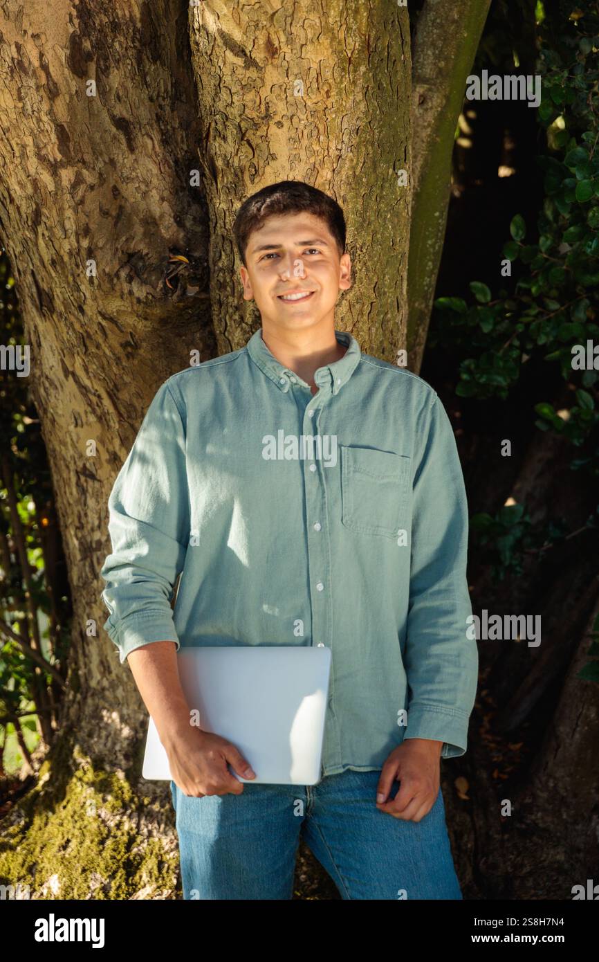 Smiling student holding laptop leaning against tree in park Stock Photo