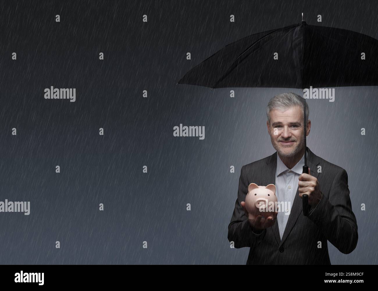 Smiling confident businessman protecting a piggy bank from the rain using an umbrella: investment protection, insurance and finance concept Stock Photo
