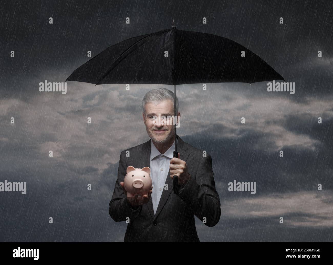 Smiling confident businessman protecting a piggy bank from the rain using an umbrella: investment protection, insurance and finance concept Stock Photo