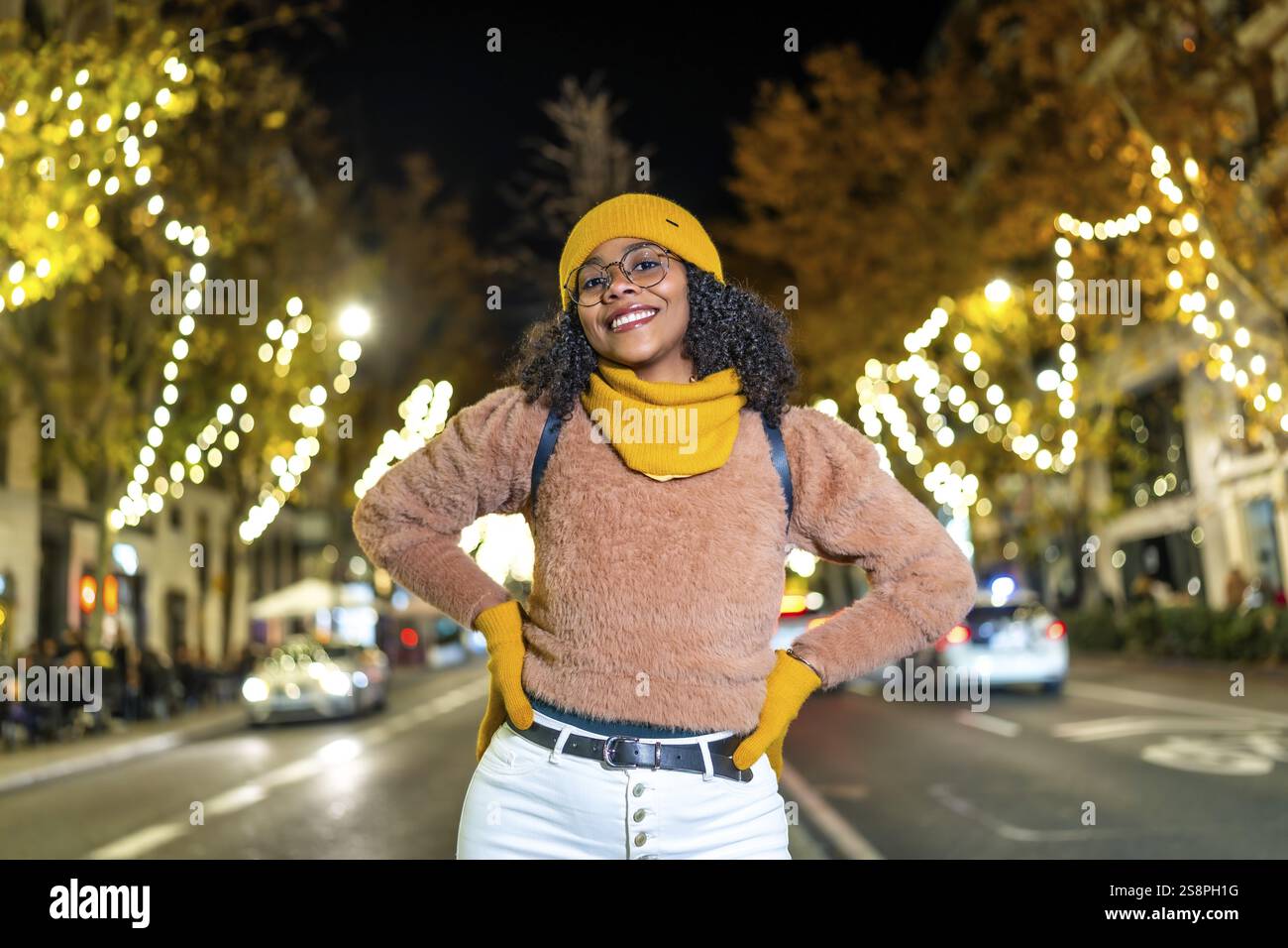 Young black woman tourist wearing yellow winter clothes and backpack, smiling with hands on her hips in a decorated city street at night Stock Photo