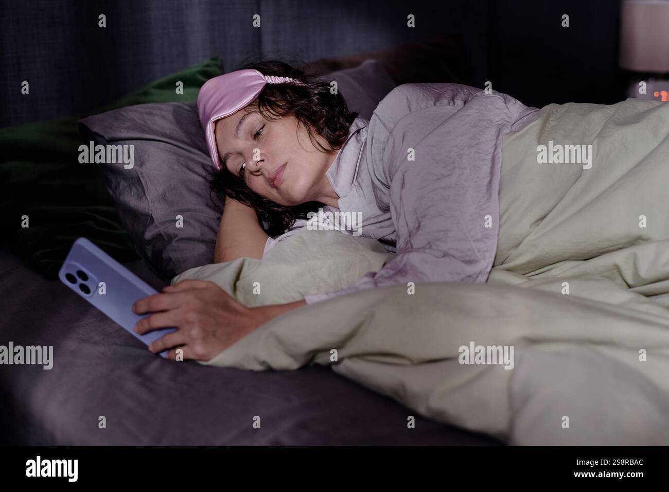 Medium close up of exhausted woman lying in bed and using smartphone while trying to fall asleep Stock Photo