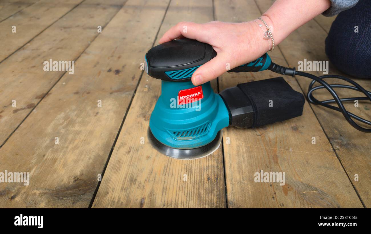 Orbital sander sanding old wooden floor. Woman in casual clothes holding sander over clean area. - Smartphone Captured Stock Image