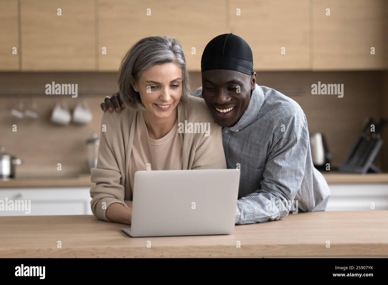Hugging mature interracial couple using notebook at home kitchen Stock Photo