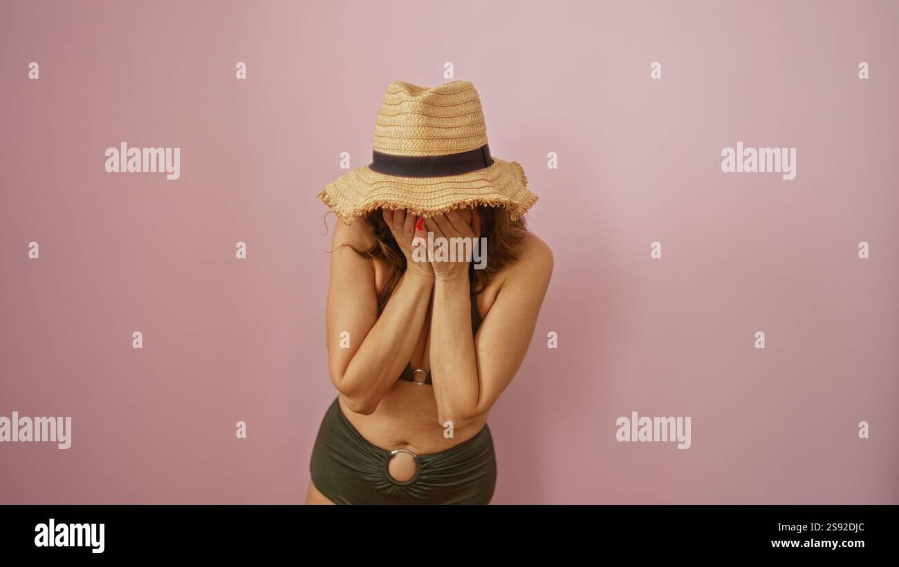 Woman covering face with hands while wearing a hat and bikini against a pink background, expressing emotions in isolation, reflecting beauty and matur Stock Photo
