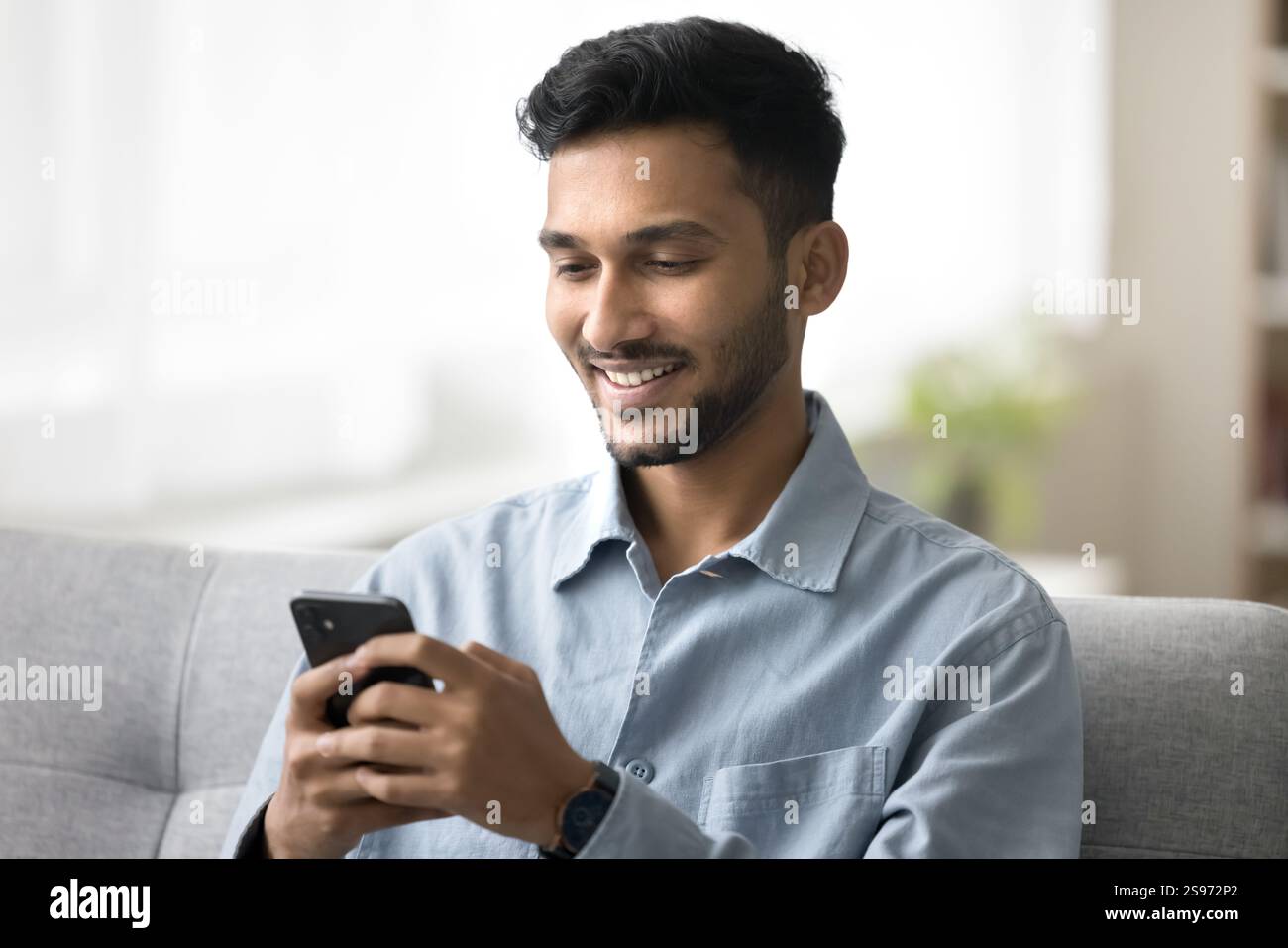 Happy young man sitting on sofa using modern mobile telephone Stock Photo