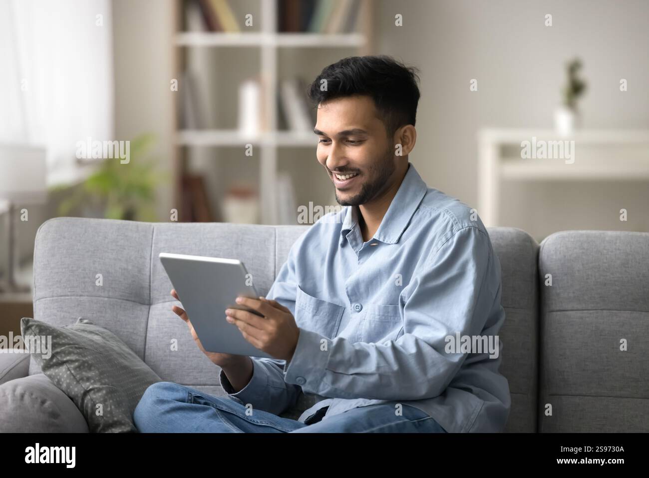 Smiling young man engaged in using pad sitting on couch Stock Photo