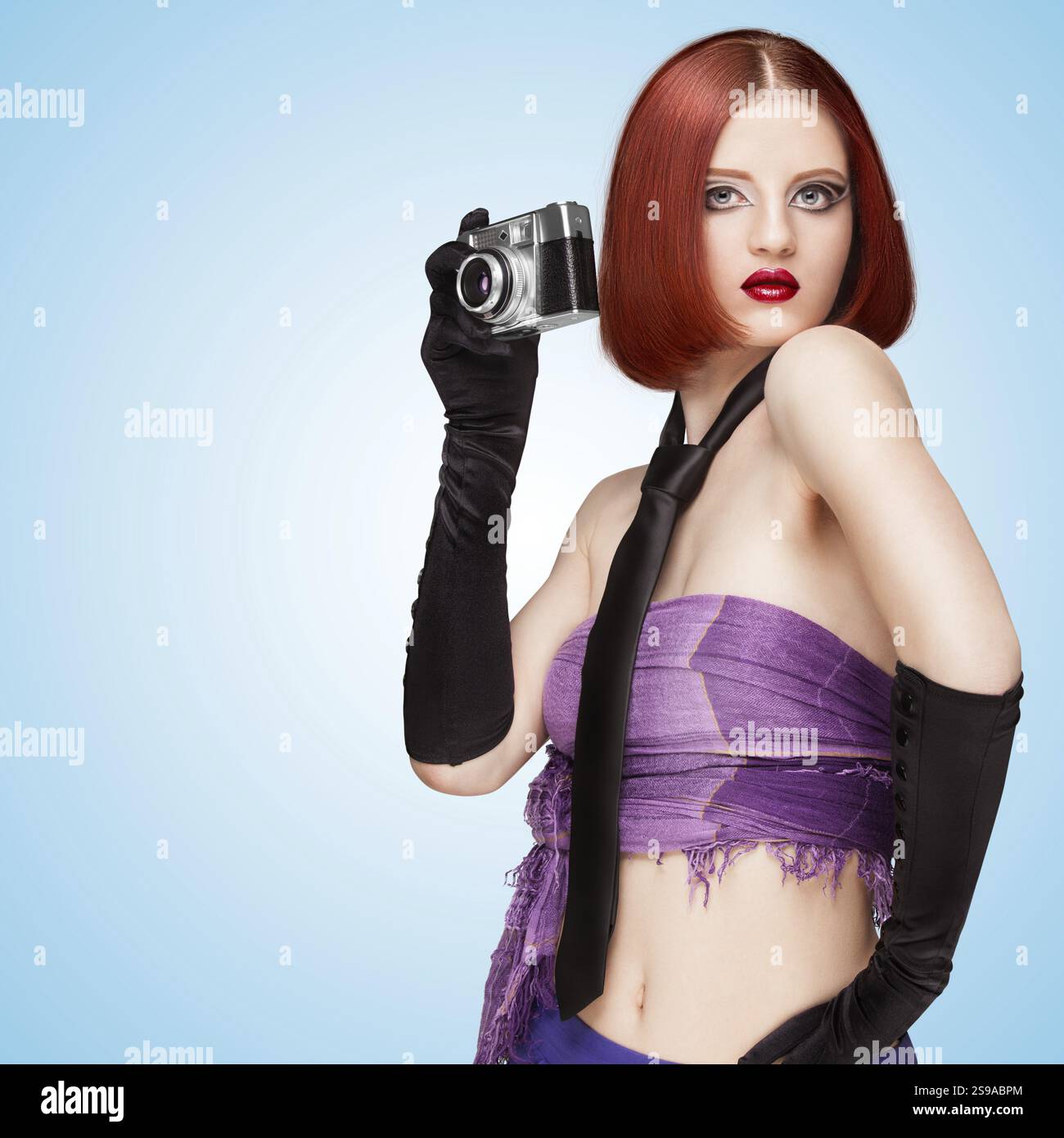 Glamorous girl in vamp style, with tie and long gloves, taking a photo with an old vintage camera on a blue background Stock Photo