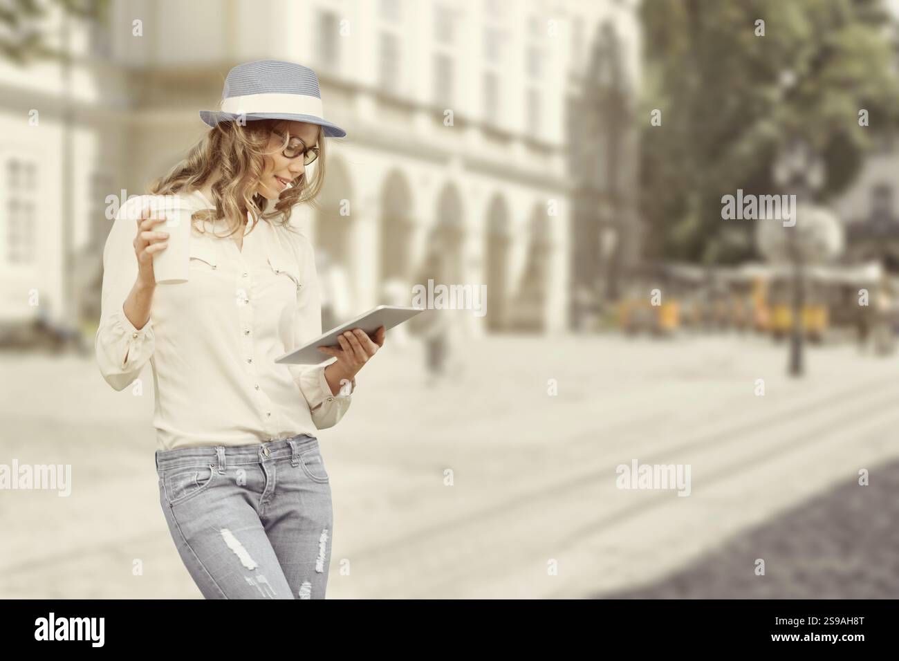 Beautiful young woman with a disposable coffee cup, holding a tablet, reading news and studying against the backdrop of an urban city Stock Photo