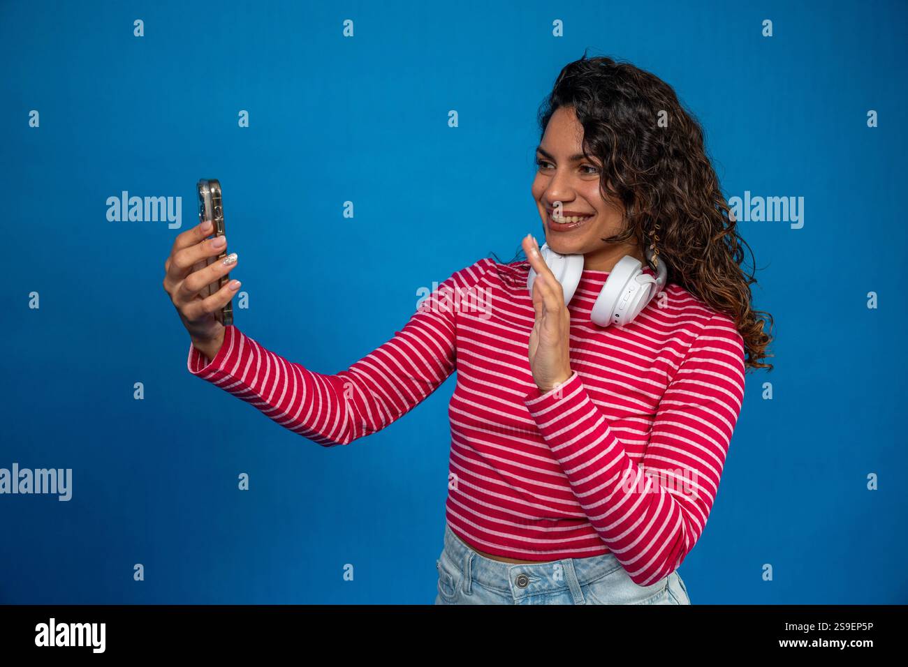 Young smiling woman with headphones making a video call with her phone waving with her hand on a blue background Stock Photo