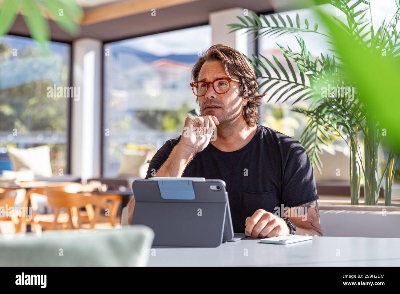 A man is sitting at a table in an office with a tablet working with a pensive expression Stock Photo