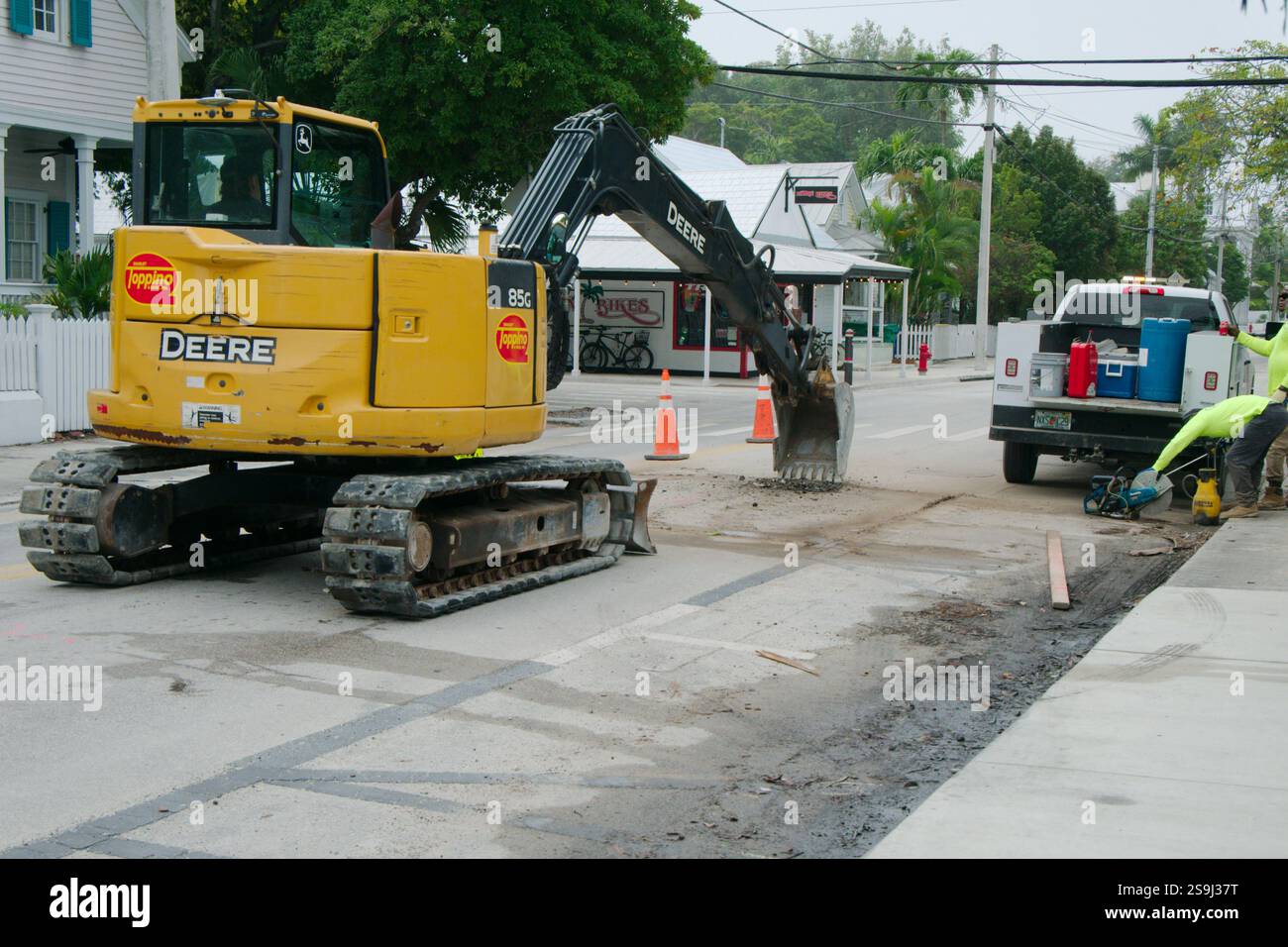 Wide view of city workers in bright yellow jackets doing street repair with a yellow backhoe bucket down on dirty roadway. Green trees and houses behi Stock Photo