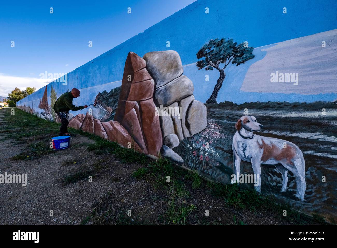 An artist paints a colourful graffiti with a rock and a dog on a wall. Arbatax Sardinia Italy FB 2024 1356 Stock Photo
