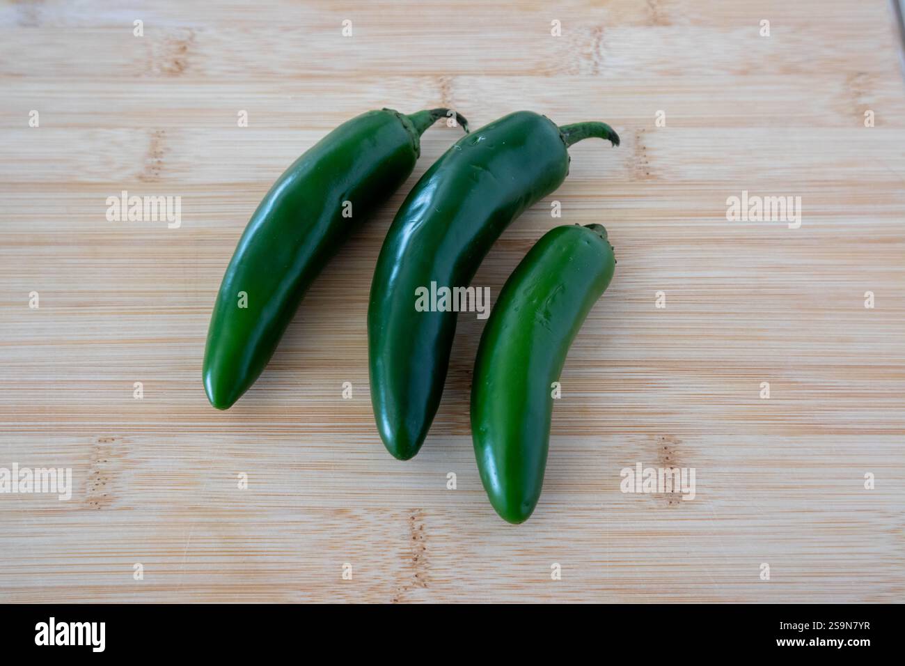 Three green jalapenos of different sizes on wood cutting board. Stock Photo