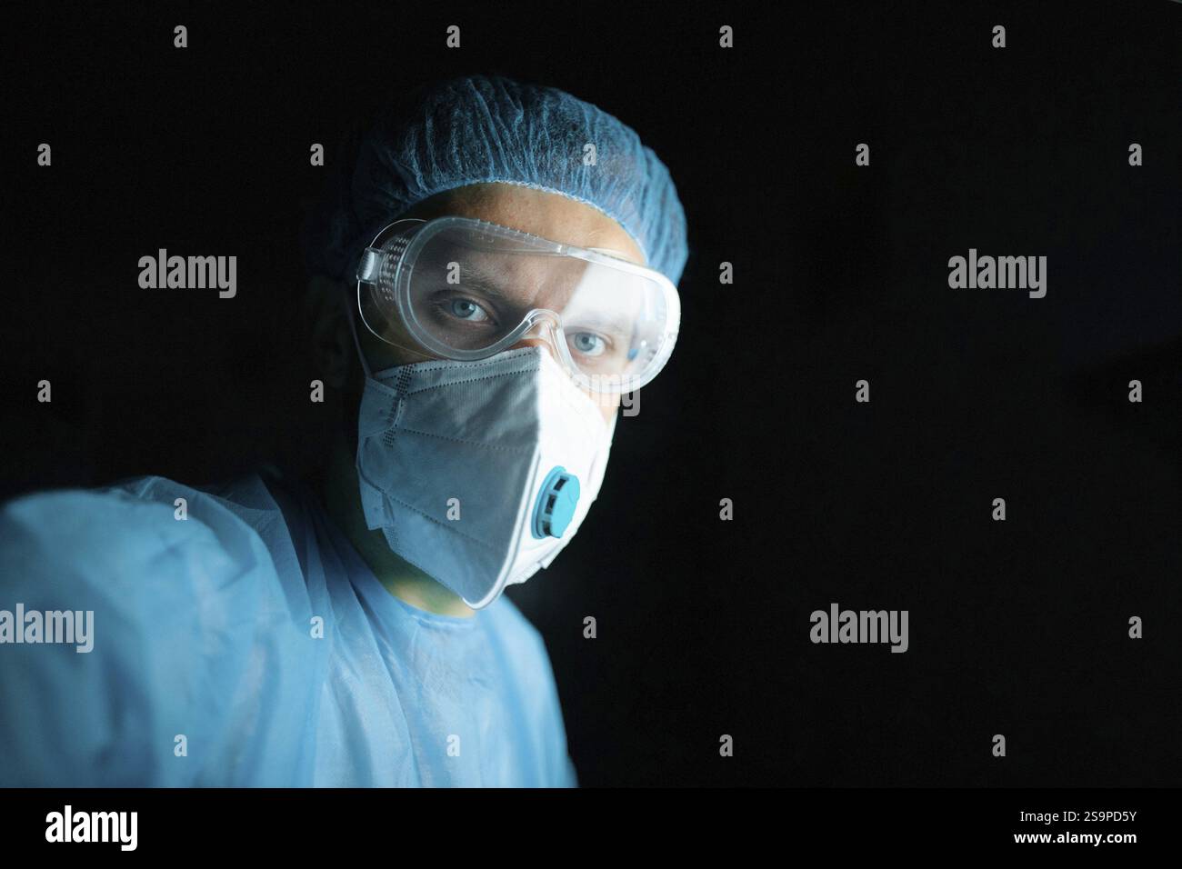 Doctor wearing goggles and mask in blue scrubs with a dark background Stock Photo