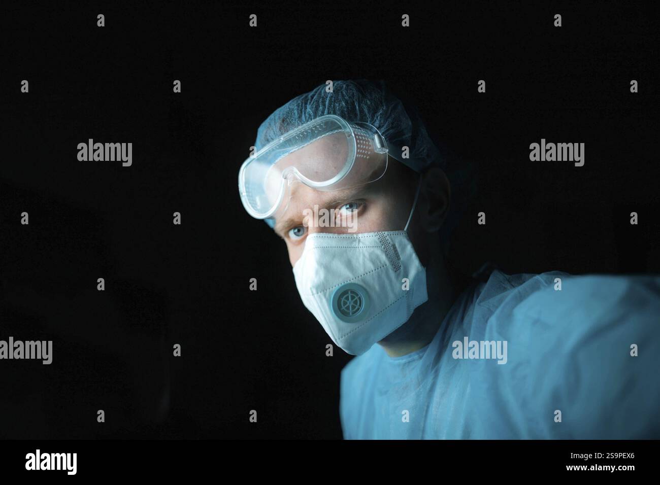 Doctor in blue scrubs wearing goggles and face mask, set against a dark backdrop Stock Photo