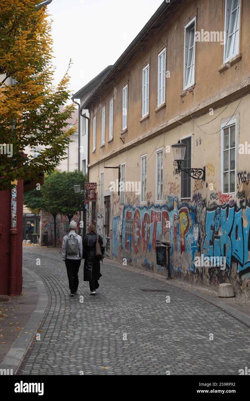 Two tourists walking down a narrow cobblestone street in Ljubljana, Slovenia, with graffiti art covering the wal Stock Photo