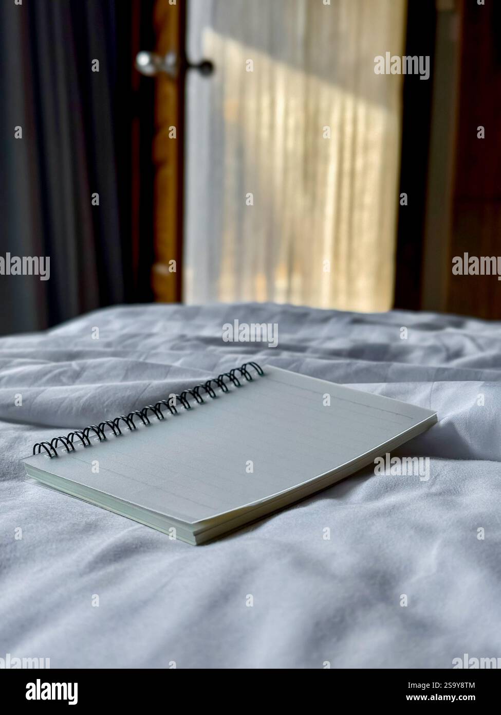 Minimalist Notebook on Bed with Natural Light A spiral notebook placed on a white bed, illuminated by soft natural light from a nearby window. Perfect - Smartphone Captured Stock Image