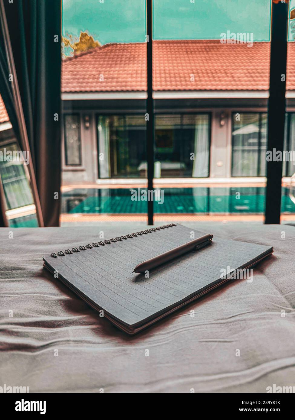 Spiral Notebook and Pen on a Bed with Blurry Pool in the Background Photo of a spiral notebook with a pen placed on the corner of a bed. In the blurry - Smartphone Captured Stock Image