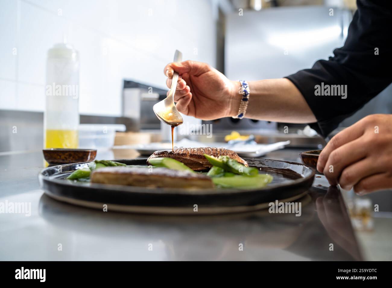 Chef plating grilled fish fillets with soy sauce and fresh vegetables in a professional kitchen, showcasing culinary precision and food artistry. Stock Photo