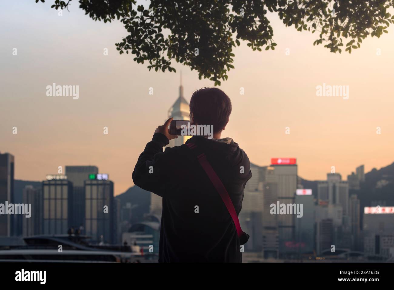 A tourist takes a photo in front of cityscape during dusk viewed from Avenue of Stars, Kowloon, Hong Kong, China, Asia Stock Photo