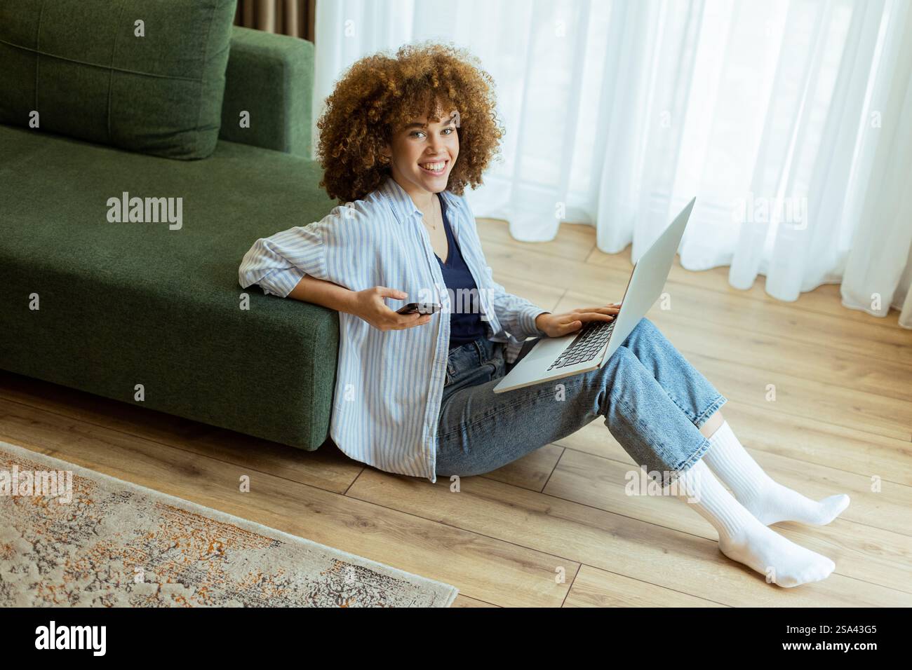 Seated comfortably on the floor, a young woman engages with her laptop while checking her smartphone. The cozy, sunlit living room creates a serene ba Stock Photo