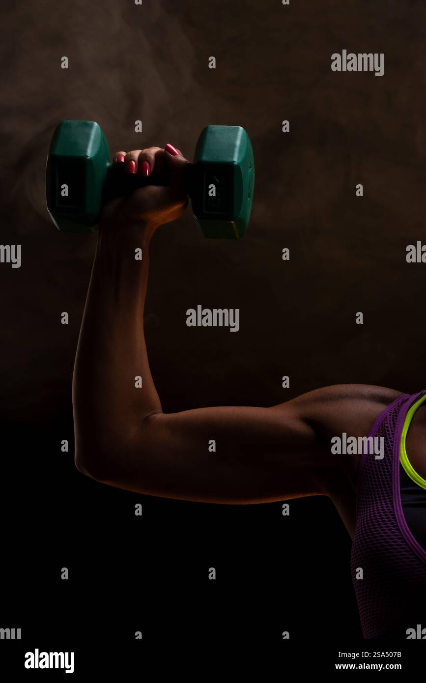 A side view of a person's arm lifting a green dumbbell, emphasizing strength and fitness. The dark background and dramatic lighting portray focus and Stock Photo