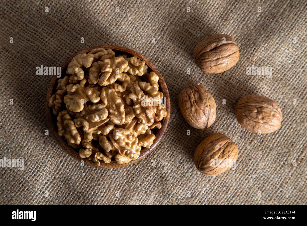 A wooden bowl filled with walnut kernels, placed alongside whole walnuts in their shells on burlap fabric Stock Photo