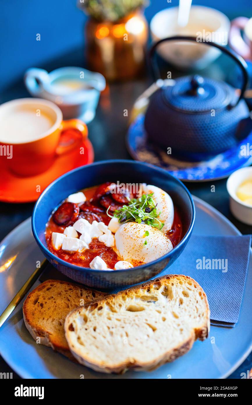 Mediterranean shakshuka served in blue bowl with poached eggs, feta cheese, microgreens, and chorizo, accompanied by sliced sourdough. Stock Photo