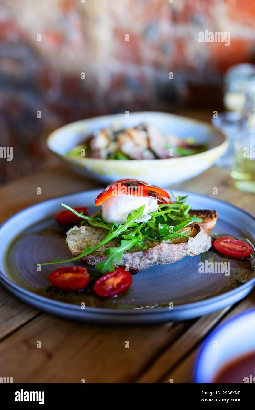 Crusty artisanal bread topped with fresh burrata cheese, peppery arugula, and roasted cherry tomatoes, served on a sleek blue ceramic plate. Stock Photo