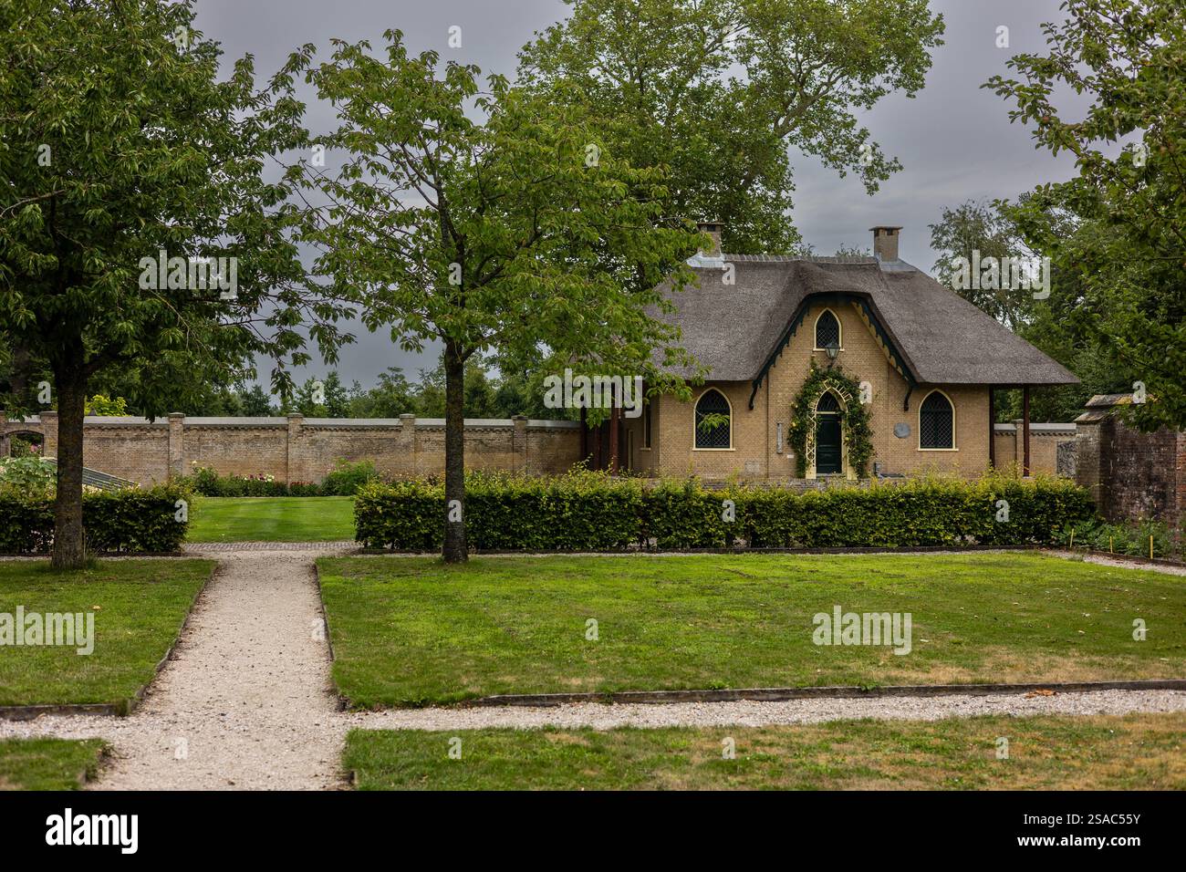 A charming brick house with a thatched roof, arched windows, and a green door covered in climbing plants. The house sits in a lush garden, with a cobb Stock Photo