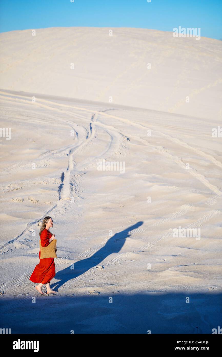 A woman in a flowing red dress carries a woven basket as she strolls across sandy dunes under the afternoon sun, casting a long shadow on the textured Stock Photo