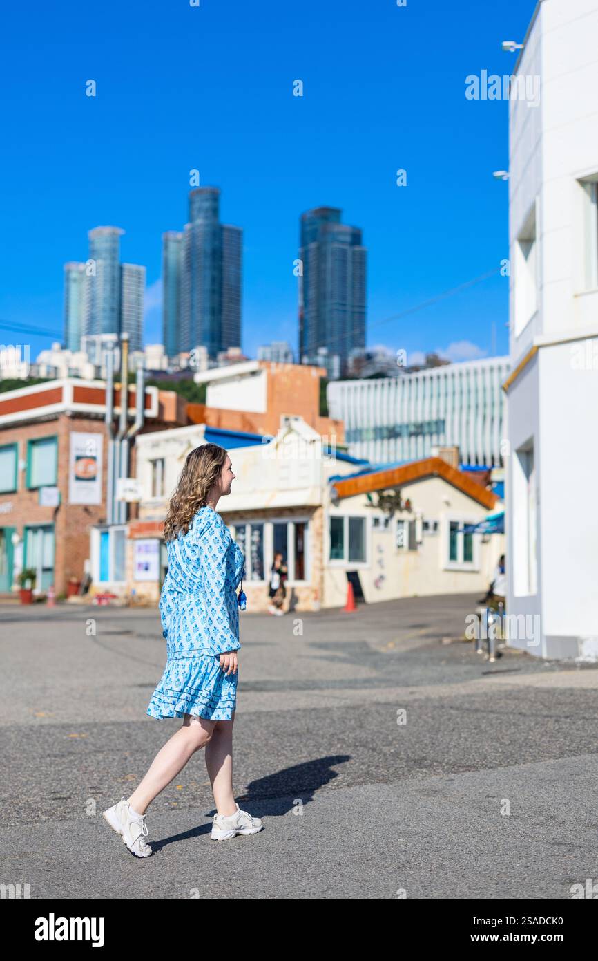 A tourist walks through a charming street in Busan, South Korea, with a mix of modern skyscrapers and colorful traditional architecture in the backgro Stock Photo