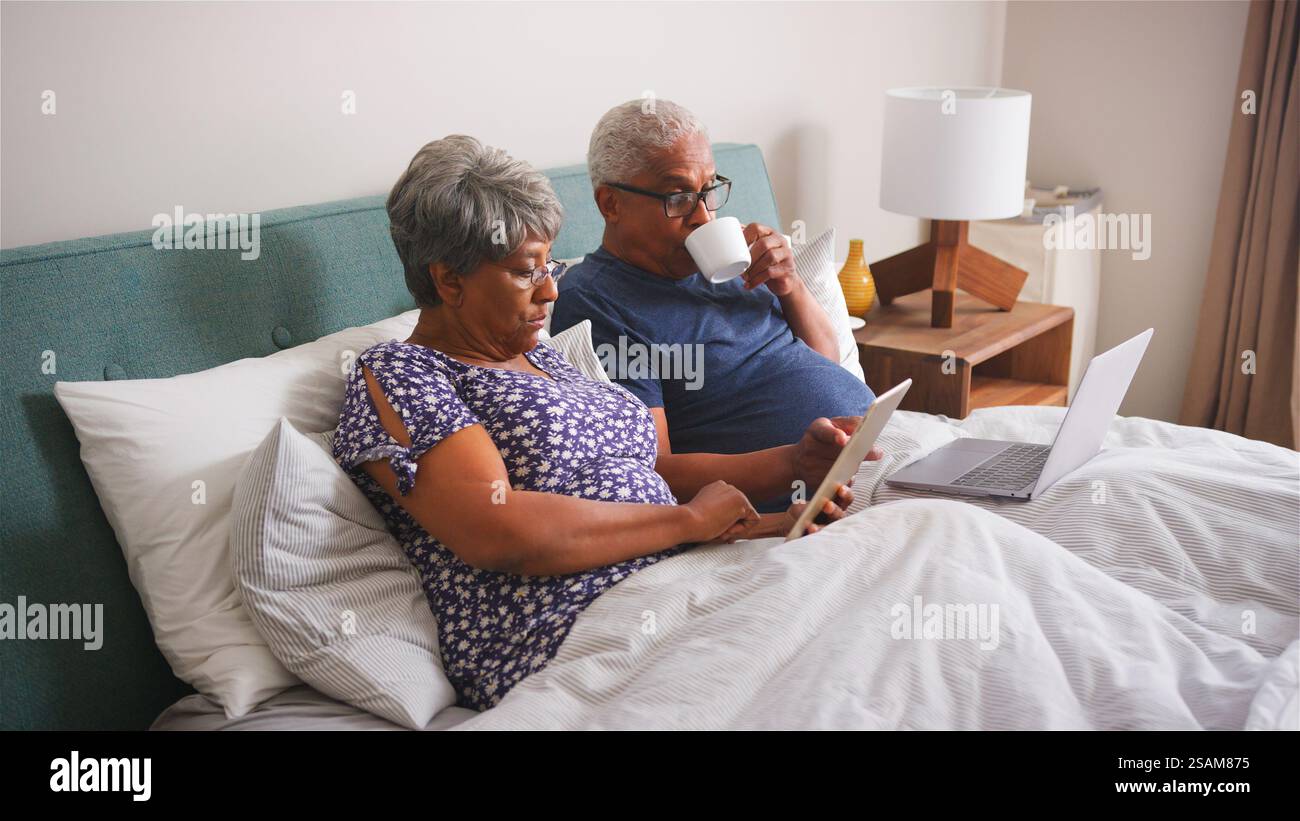 Senior Couple Lying In Bed At Home Using Digital Devices Stock Photo
