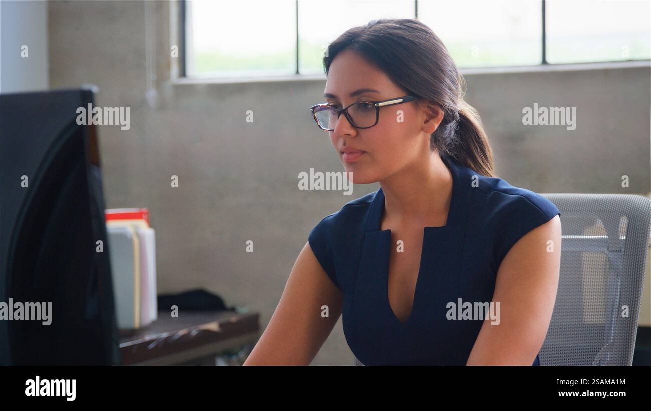 Businesswoman Wearing Glasses Sitting At Desk In Office Working On Computer Stock Photo