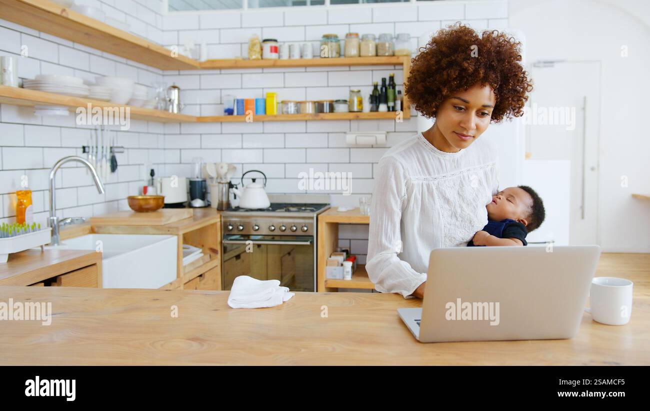 Multi-Tasking Mother Holds Sleeping Baby Son Whilst Working On Laptop In Kitchen Stock Photo