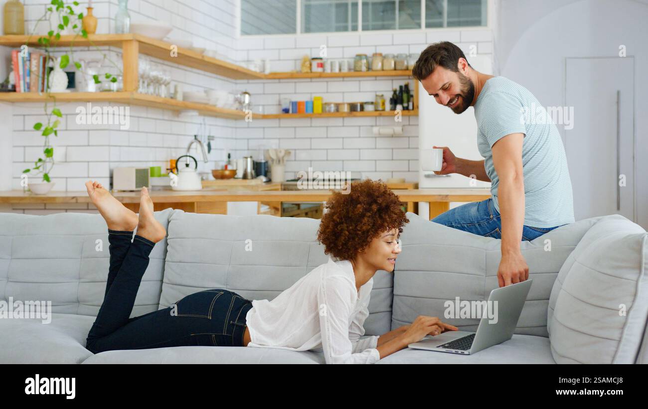 Young Couple At Home Woman On Sofa Using Laptop Computer With Man With Hot Drink In Kitchen Behind Stock Photo