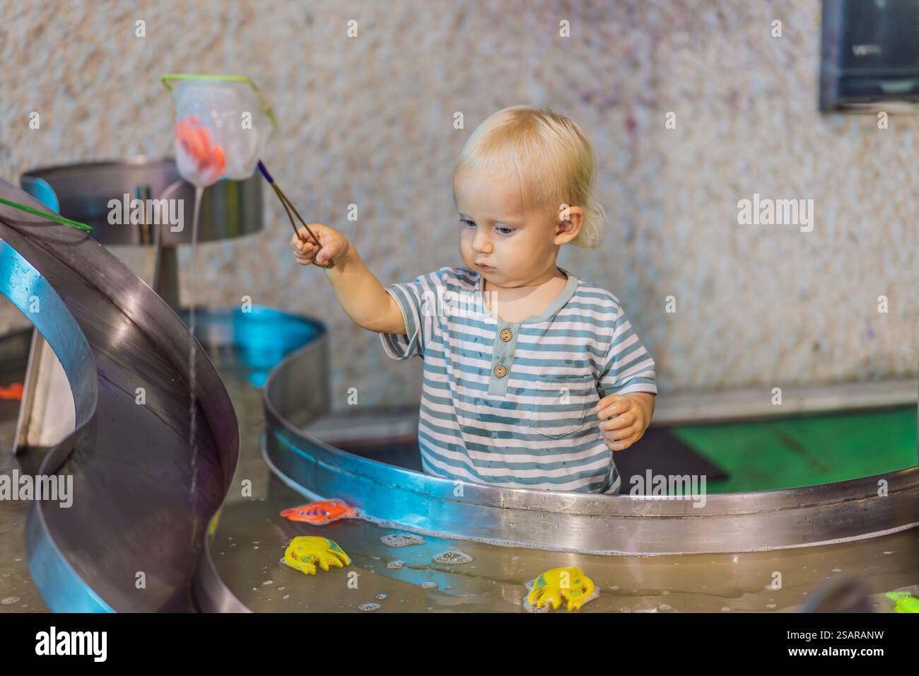 Toddler boy catching toy fish with a net in a vibrant water play area ...