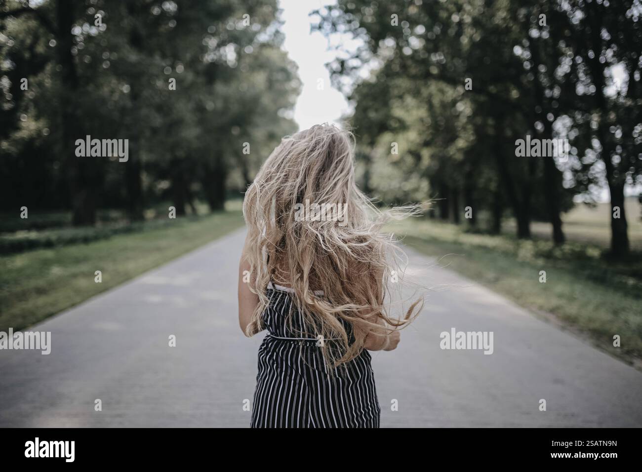 A girl with long blonde hair walks on a tree-lined road in a striped ...