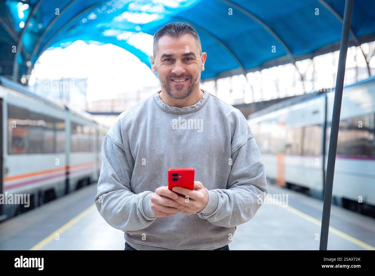 Smiling Man Using Red Smartphone at Train Station looking at camera Stock Photo