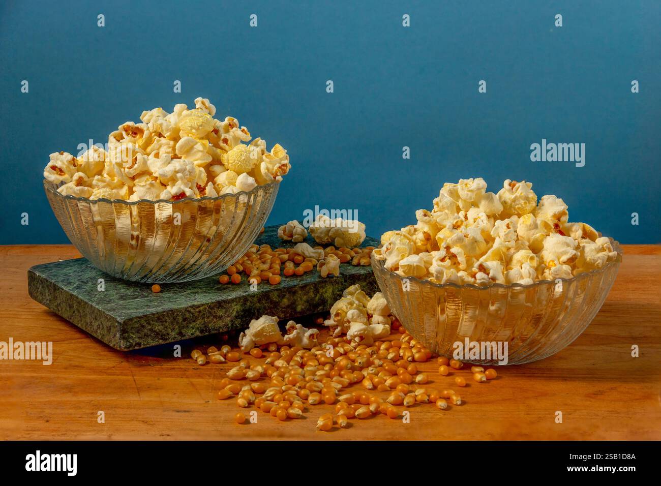 Two glass bowls filled with freshly popped popcorn surrounded by scattered corn kernels on a rustic wooden table, featuring a blue background. Stock Photo
