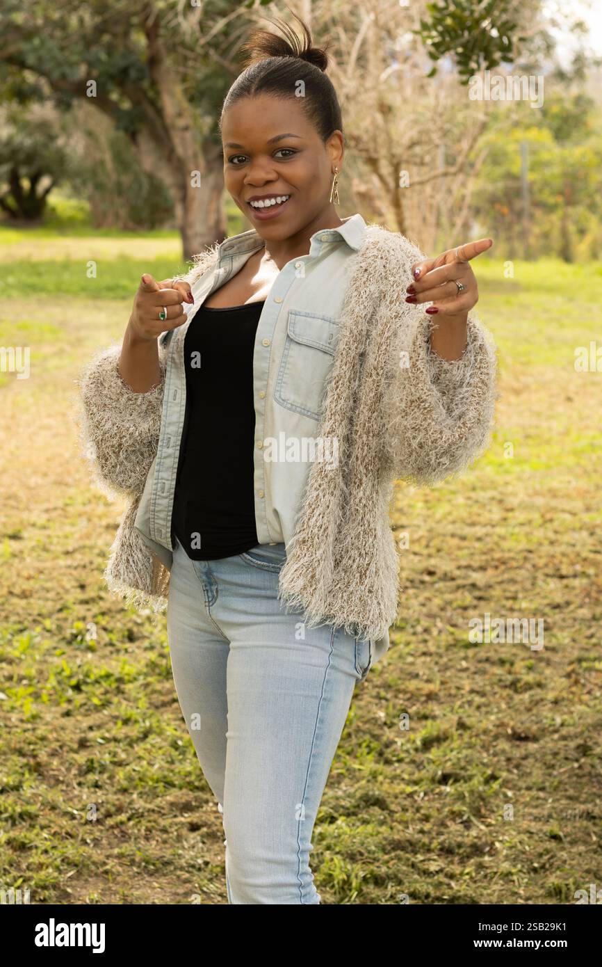 A black woman is standing outdoors, smiling confidently while pointing with both hands. Stock Photo