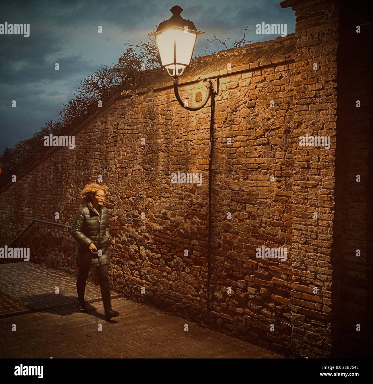 A woman in a warm jacket walks down the alley. The streetlamp’s glow highlights her face, while her shadow stretches along the wall, telling a story - Smartphone Captured Stock Image